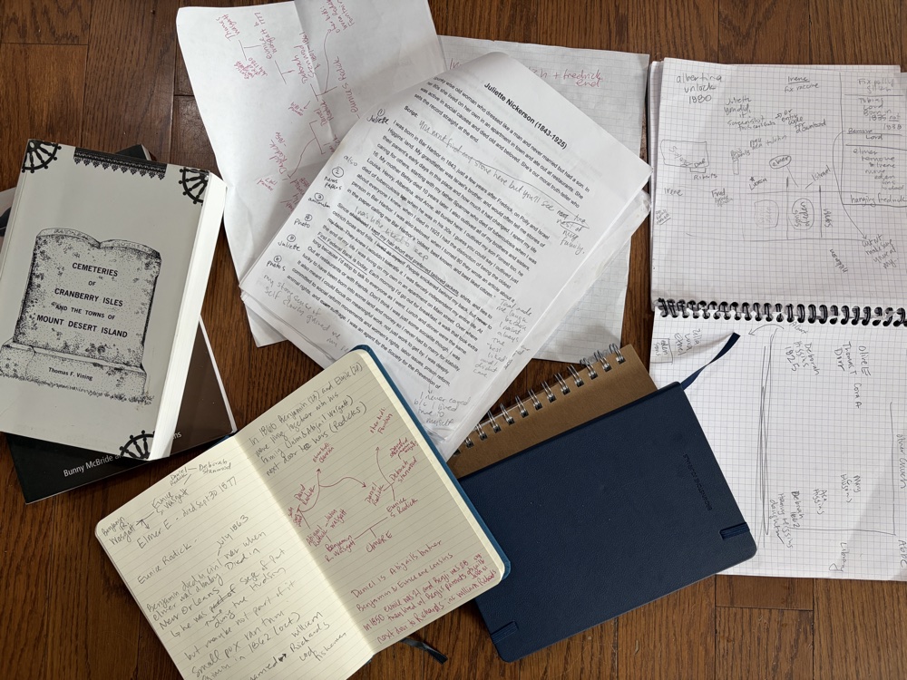 A photograph of physical research materials: open notebooks with handwritten notes in pencil and red ink, marked-up printed pages, family-tree diagrams on graph paper, and the spiral-bound book Cemeteries of Cranberry Isles and the Outer Mount Desert Island spread across a wood floor.