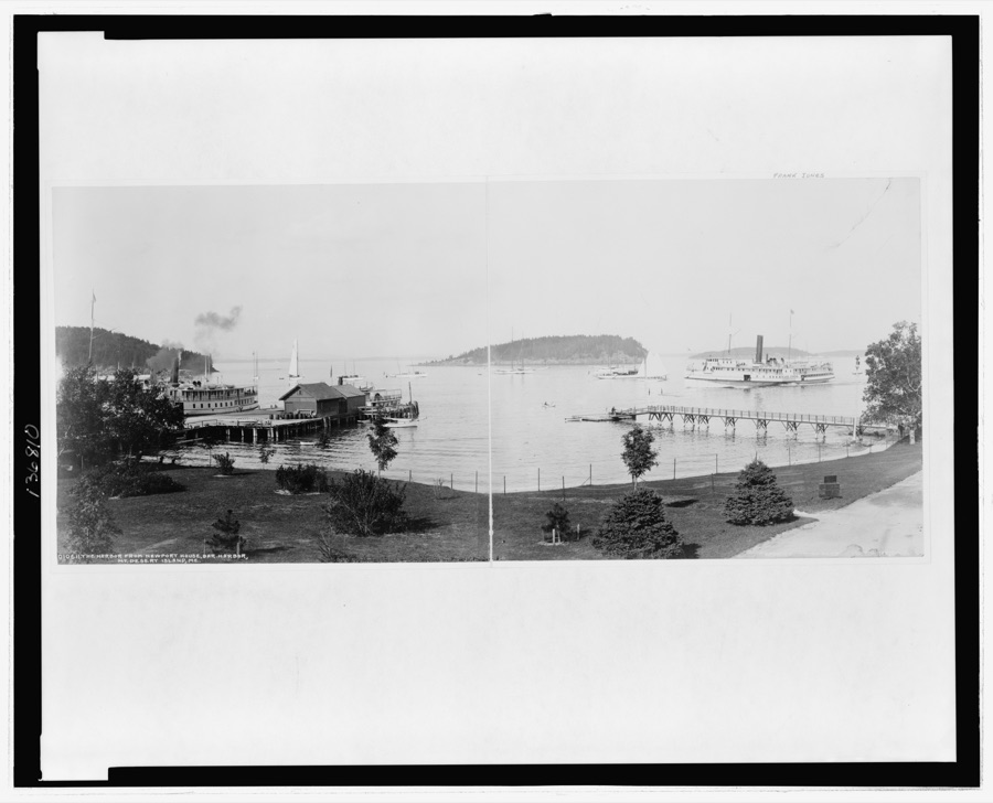 A panoramic view of the harbor at Bar Harbor seen from the grounds of the Newport House, with two steamboats tied up at the piers and Bar Island in the distance, 1901.